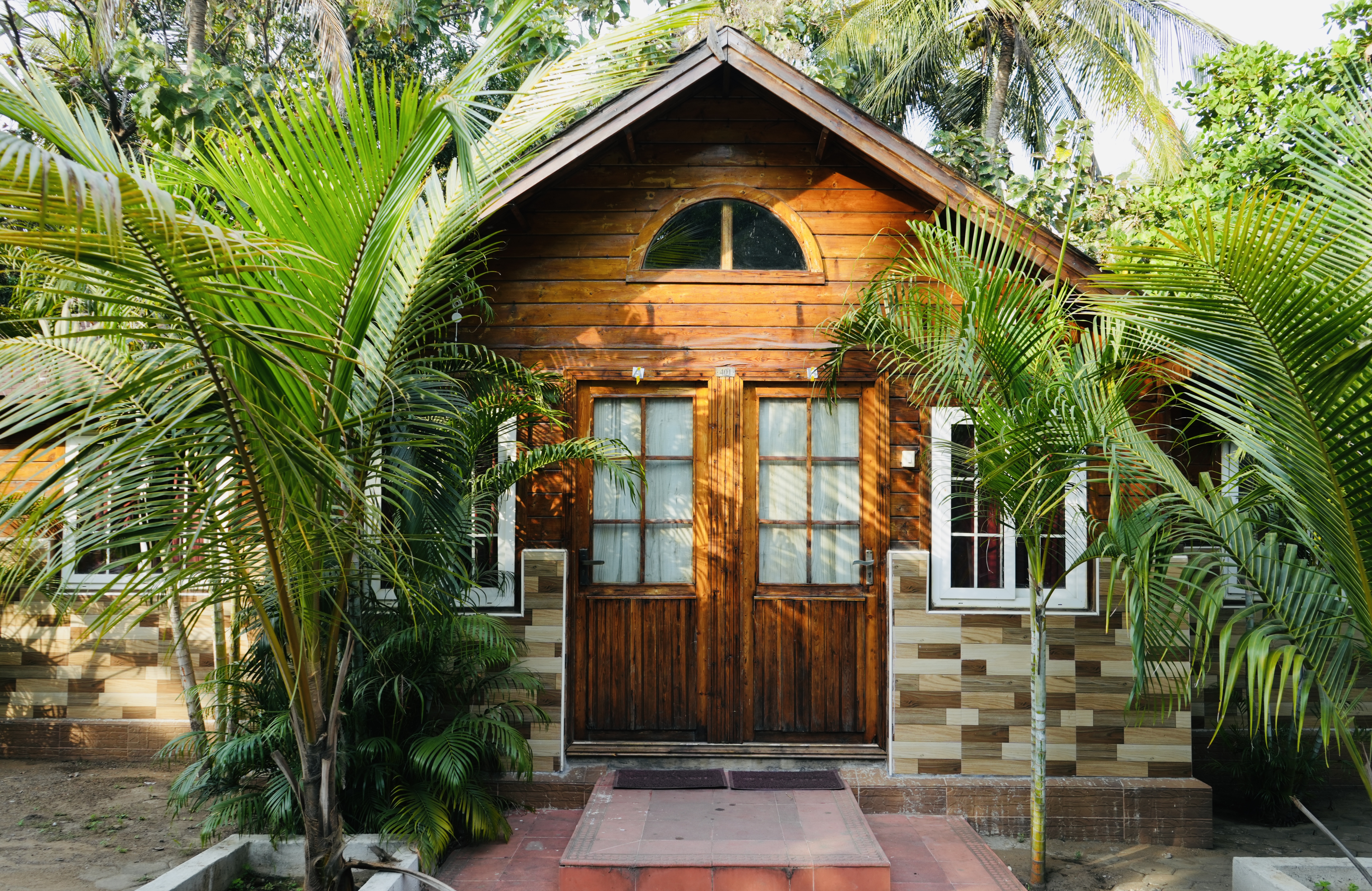 Exterior view of a premium wooden cottage at Blue Bay Beach Resort, Mahabalipuram, surrounded by lush palm trees and tropical greenery.