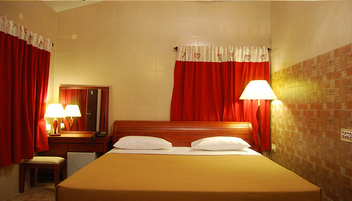 Bed with a night lamp by its right side and a dressing table with a mirror and a chair with a table lamp along with an intercom in Bluebay beach resort, Chennai